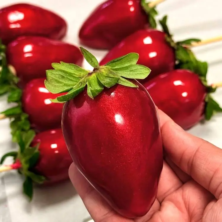 Uma mão segura um morango vermelho brilhante e caramelizado, com suas folhas verdes intactas e um palito inserido na base. Ao fundo, outros morangos caramelizados semelhantes aparecem ligeiramente desfocados, sugerindo uma bandeja cheia dessas guloseimas.