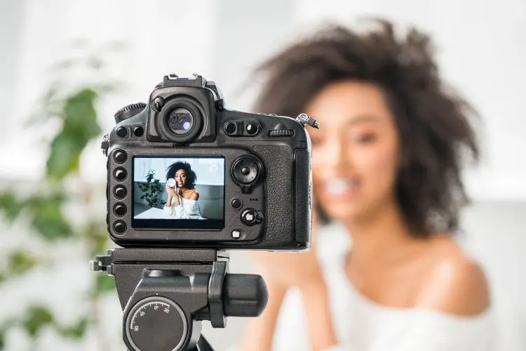 A imagem mostra uma câmera DSLR focada em uma mulher sorridente, com cabelo cacheado, ao fundo. Ela está sentada, usando uma blusa clara. No visor da câmera, sua imagem aparece nítida. O ambiente é claro, com plantas ao fundo.