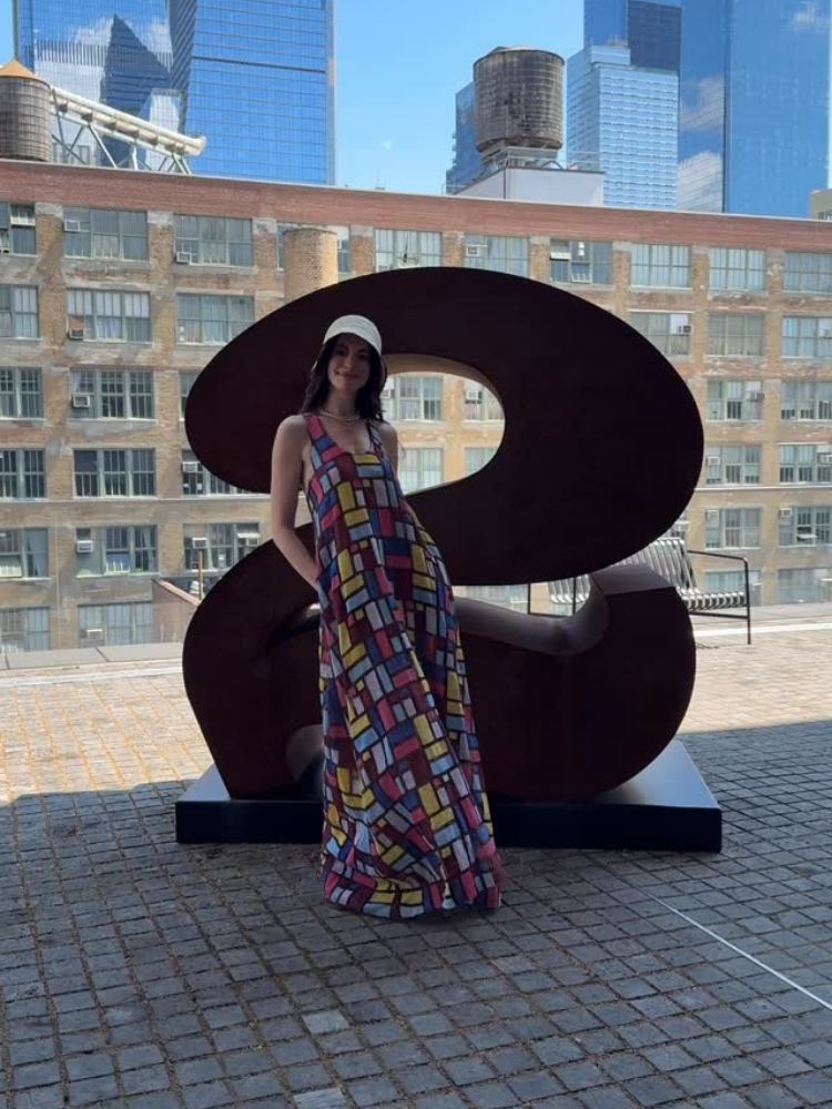 Anne Hathaway em frente a uma escultura de "2" usando vestido colorido de patchwork, bucket branco 