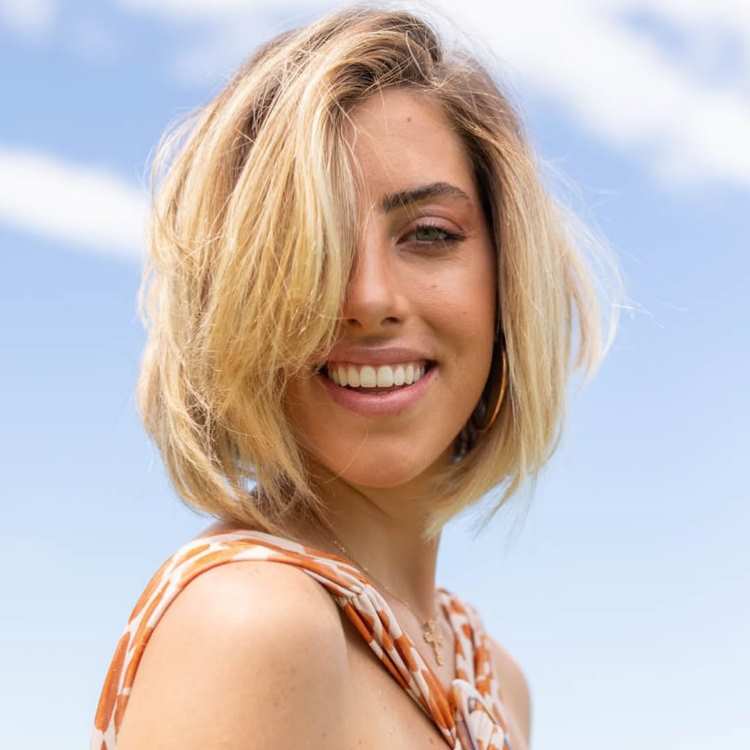 Mulher sorridente com cabelo curto estilo bob loiro despojado, usando roupa com estampa laranja e branca, brincos dourados e colar delicado. Está ao ar livre sob céu azul com nuvens leves, transmitindo uma expressão alegre e confiante.