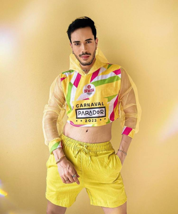 A imagem apresenta um homem usando um abadá customizado amarelo com detalhes em rosa, verde e branco, estampado com "Devassa", "Carnaval Parador 2023". A peça possui mangas transparentes e um design cropped, combinando com shorts amarelos em um fundo monocromático.