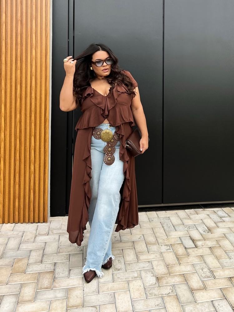 Mulher de pele clara com blusa marrom de babados, maxi cinto de couro e calça jeans 