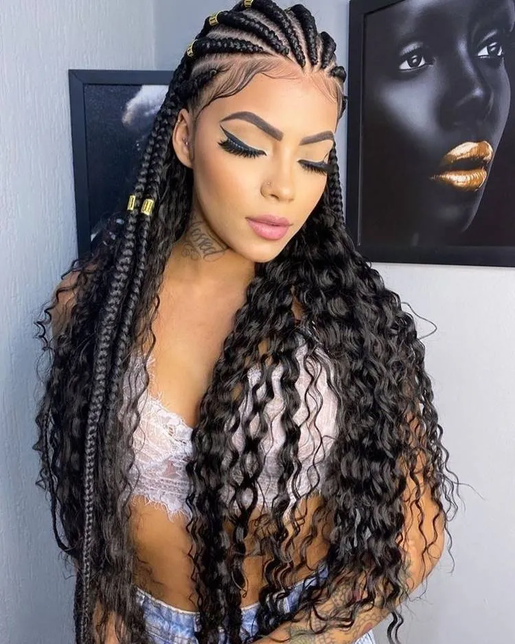 Mulher com top de renda e shorts jeans, cabelos longos cacheados, usa tranças nagô com joias no cabelo. Fundo parede com quadros de pessoas negras usando batom dourado