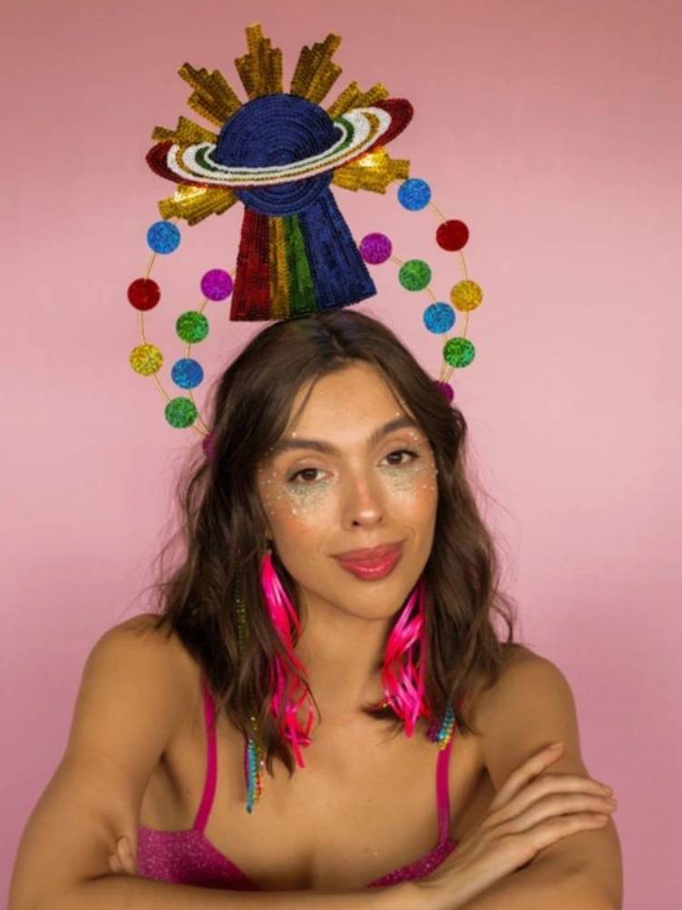 Mulher de pele clara e cabelo castanho usando brinco de fita rosa e tiara de planeta com arco-iris, anéis e outras bolinhas coloridas.