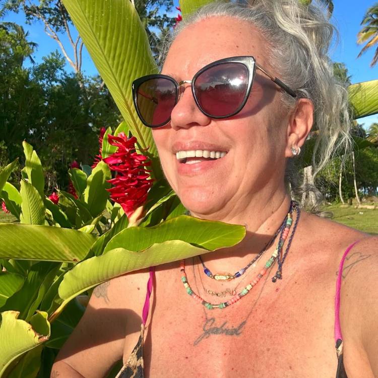 Astrid Fontenelle de biquíni ao lado de plantas, com sua tatuagem em homenagem ao filho, um cordão escrito "Gabriel"
