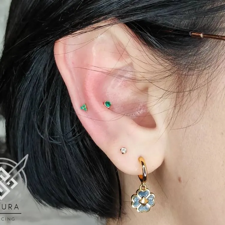 Orelha com piercing com pedra verde no snug e dois brincos no lóbulo, sendo um ponto de luz e outro uma argola dourada ocm flor azul