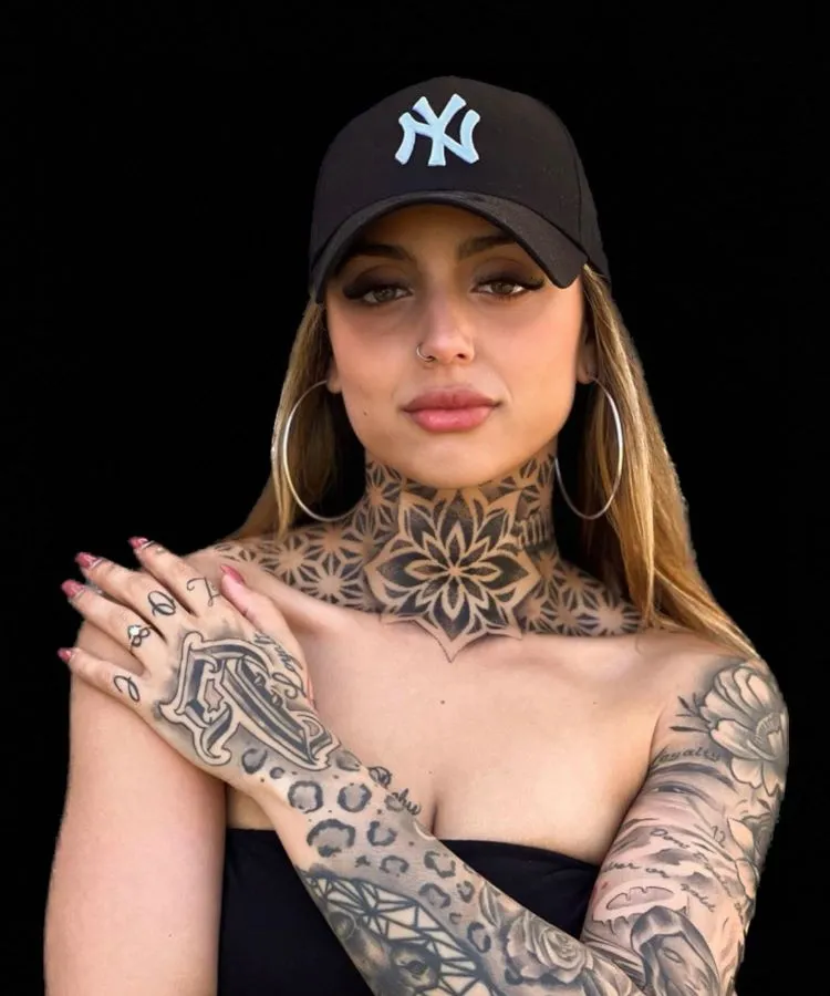 A imagem mostra uma pessoa de frente com cabelo loiro, usando um boné preto e brincos grandes. Ela tem uma tatuagem no pescoço de um grande padrão geométrico em preto, que se estende até o peito. O braço direito também está coberto por tatuagens intricadas.
