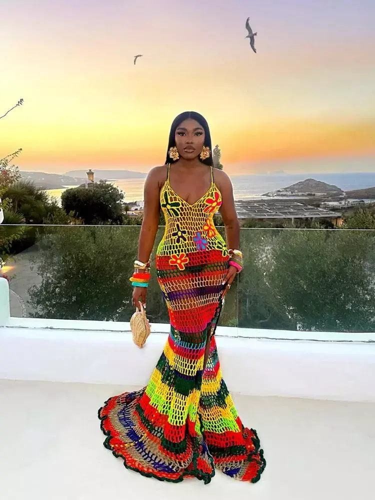 Mulher de pele negra em prédio alto com mar e pôr do sol ao fundo usando vestido longo de crochê colorido