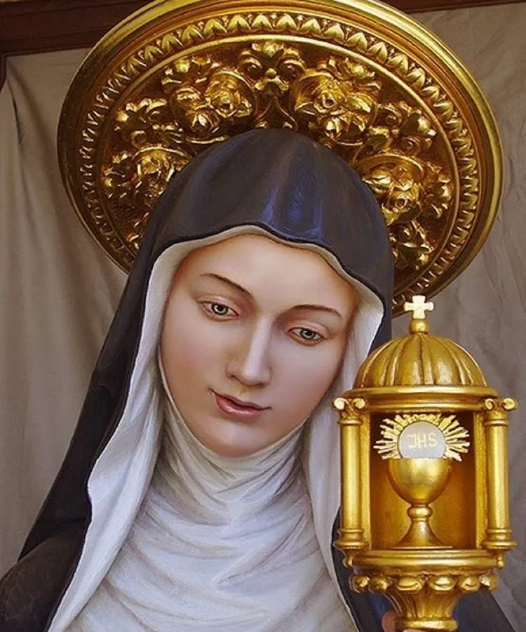 A imagem mostra Santa Clara de Assis com um hábito marrom e véu preto. Ela segura um ostensório dourado com uma hóstia branca. Santa Clara tem um rosto sereno e usa uma auréola dourada decorada com flores. O fundo é neutro, destacando a figura da santa.


