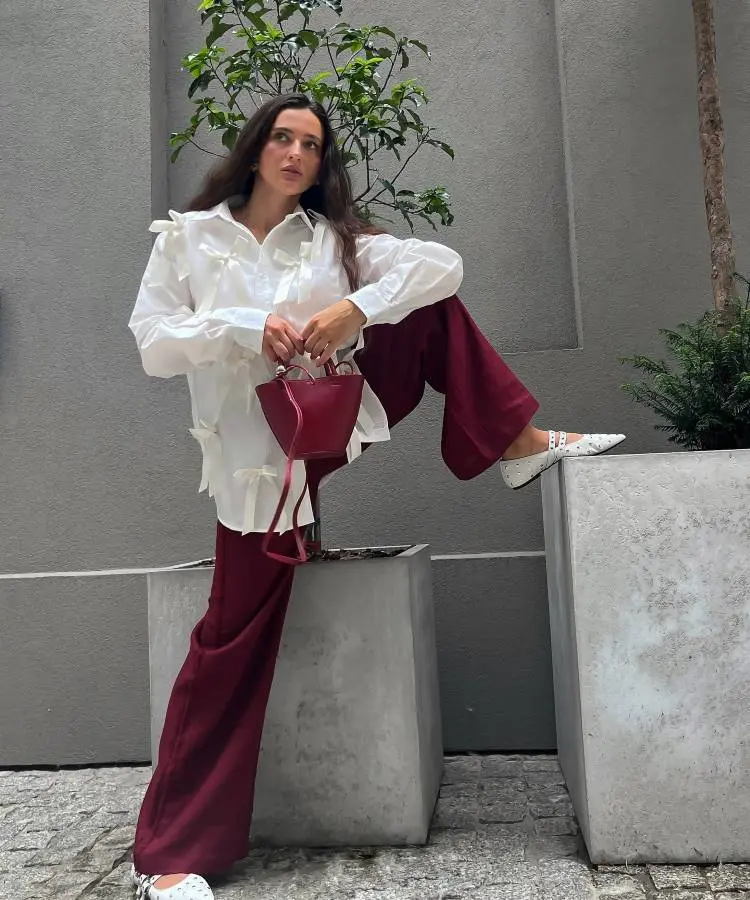 Uma mulher posa com uma perna apoiada em um vaso de concreto. Ela usa uma blusa branca com laços, calças largas de cor vinho, sapatos brancos estampados e segura uma pequena bolsa vermelha.