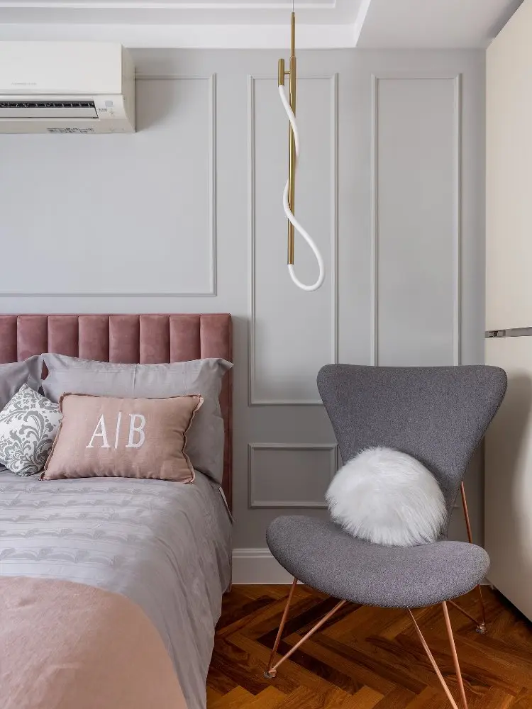 Parte de quarto com parte de cama com cabeceira rosa, poltrona cinza, lustre e parede branca com  moldura 