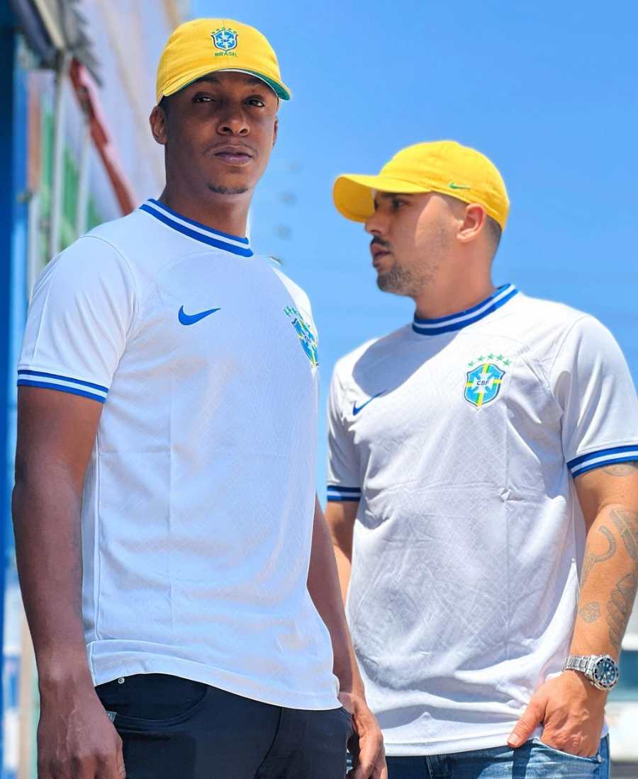Imagem com fundo de céu azul. No centro, 2 homens usando camiseta branca do Brasil + boné amarelo.