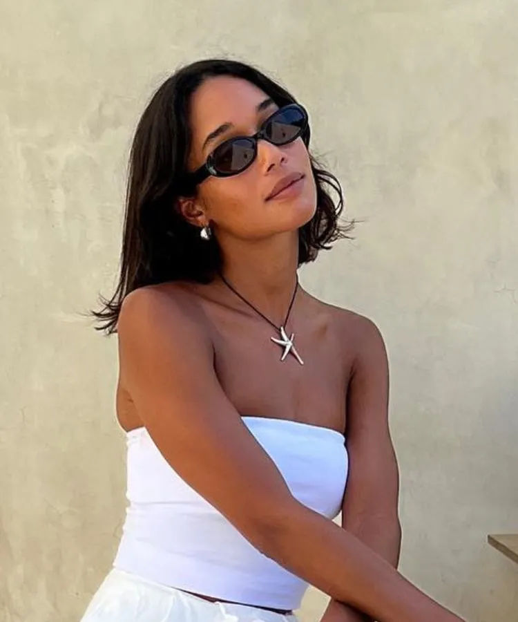 A imagem mostra uma mulher com um corte de cabelo bob castanho na altura do ombro, usando óculos de sol ovais escuros. Ela veste um top tomara que caia branco e um colar preto com um pingente prateado em forma de estrela do mar. Brincos pequenos e prateados complementam o visual.







