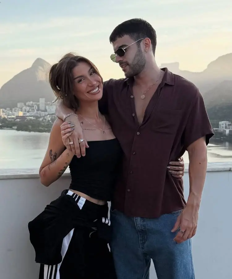 Casal Bianca Andrade e Diego Cruz posa em um terraço com vista para uma cidade e montanhas ao pôr do sol. A mulher sorri para a câmera enquanto o homem a abraça, olhando para ela.