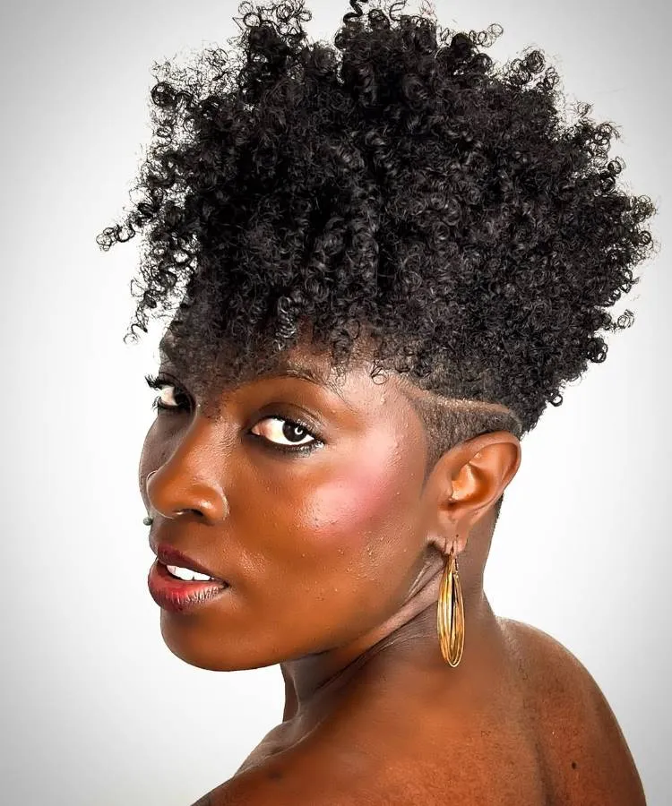 Uma mulher com um corte de cabelo cacheado em camadas, raspado nas laterais com um desenho. Ela usa grandes brincos de argola e olha para a câmera com maquiagem destacada nos olhos e lábios.