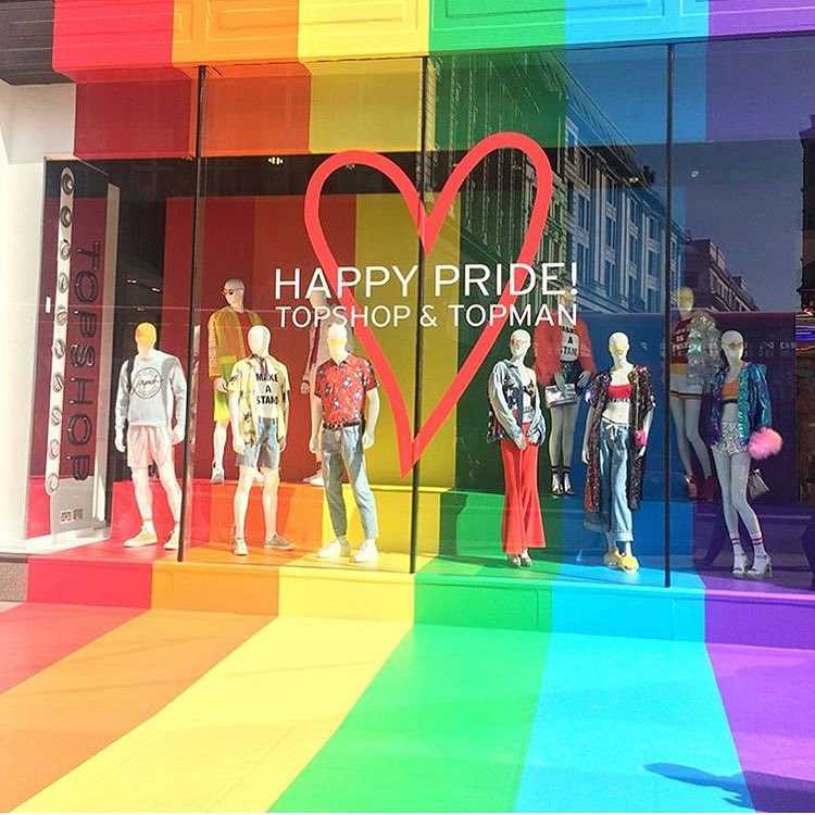 Vitrine da loja Topshop & Topman com fachada em tema arco-íris, destacando a frase "Happy Pride!" e um coração vermelho. Manequins expõem roupas coloridas e estilosas. Piso e fundo vibrantes celebram o mês do Orgulho LGBTQIA+, promovendo diversidade e inclusão.