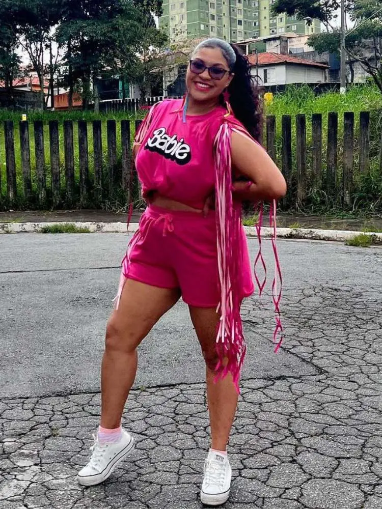 Mulher de pele negra com blusa rosa da Barbue, shorts rosa e ombreira rosa de franjas
