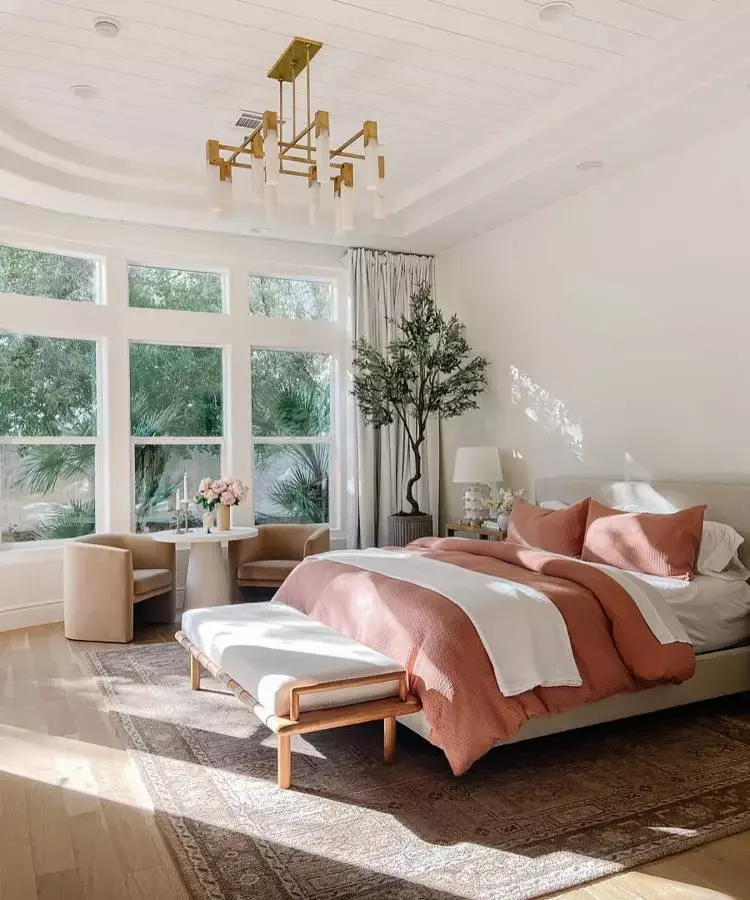 Quarto de casal com cama grande nas cores branco e salmon. Planta ao lado, parede de vidro mesa e poltronas 