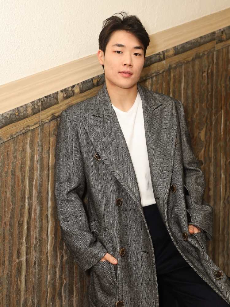 Arthur Paek usando blusa branca, sobretudo cinza e calça preta