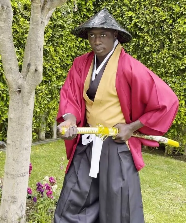 Khaby Lame veste um quimono vermelho e amarelo, com calças pretas largas e um chapéu cônico asiático. Ele segura uma espada dourada com as duas mãos, em um jardim verde.