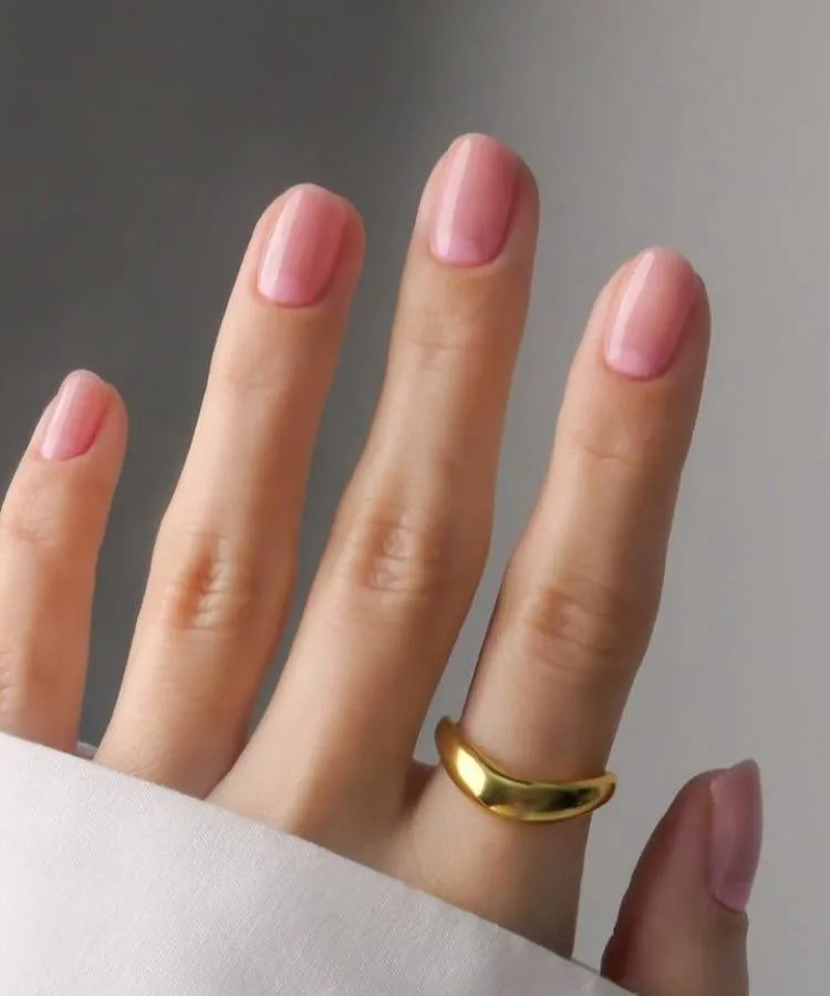 Unhas elegantes, curtas e simples. Transparente na cor rosinha. A mão possui um anel dourado no dedo indicador.
