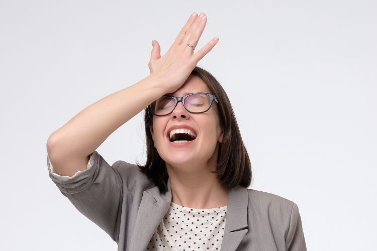Uma mulher, usando óculos e uma jaqueta cinza, está com a mão na testa, parecendo expressar frustração ou esquecimento. Ela tem a boca aberta em uma expressão de surpresa ou exasperação, contra um fundo branco simples.







