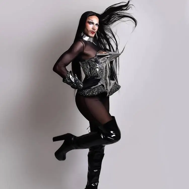 Drag Queen com corset prateado futurista, blusa transparente e meia-calça preta. Botas de cano alto em verniz completam o visual glamuroso. Cabelo liso e longo solto, movimento captado na pose confiante, transmitindo força, brilho e teatralidade típica de apresentações drag.