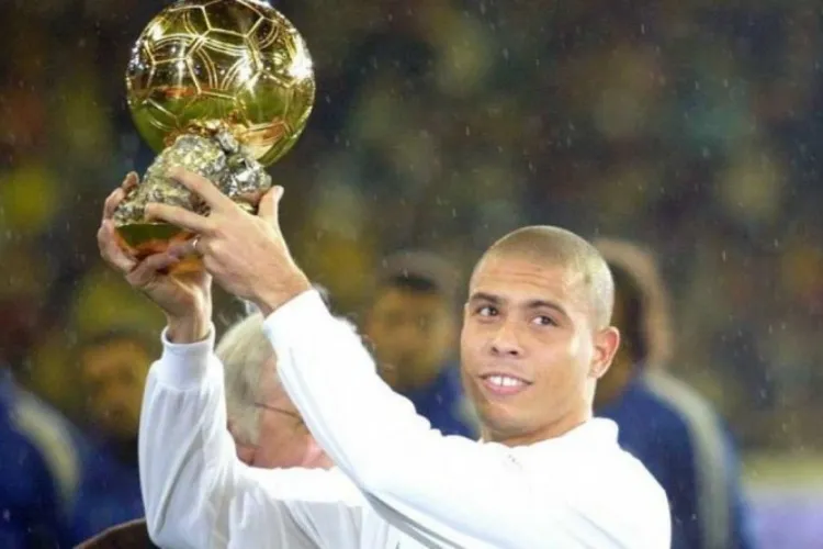 A imagem retrata um homem segurando e erguendo o prêmio Bola de Ouro, sorrindo. Ele veste roupa branca, tem cabelo raspado e está em um ambiente externo à noite, com público desfocado ao fundo e luzes ao redor.