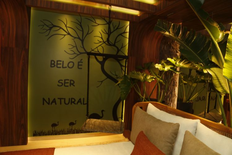 quarto com cama, almofadas em bege e laranja, parede verde escrito "belo é ser natural" e balanço de cordas com estofado marrom