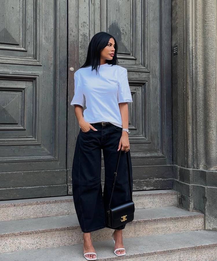A imagem destaca uma calça barrel leg preta, tendência jeans 2025, com cintura alta e silhueta cilíndrica ampla. O look é elegante e moderno, combinado com blusa branca minimalista, sandálias de salto e bolsa preta sofisticada.