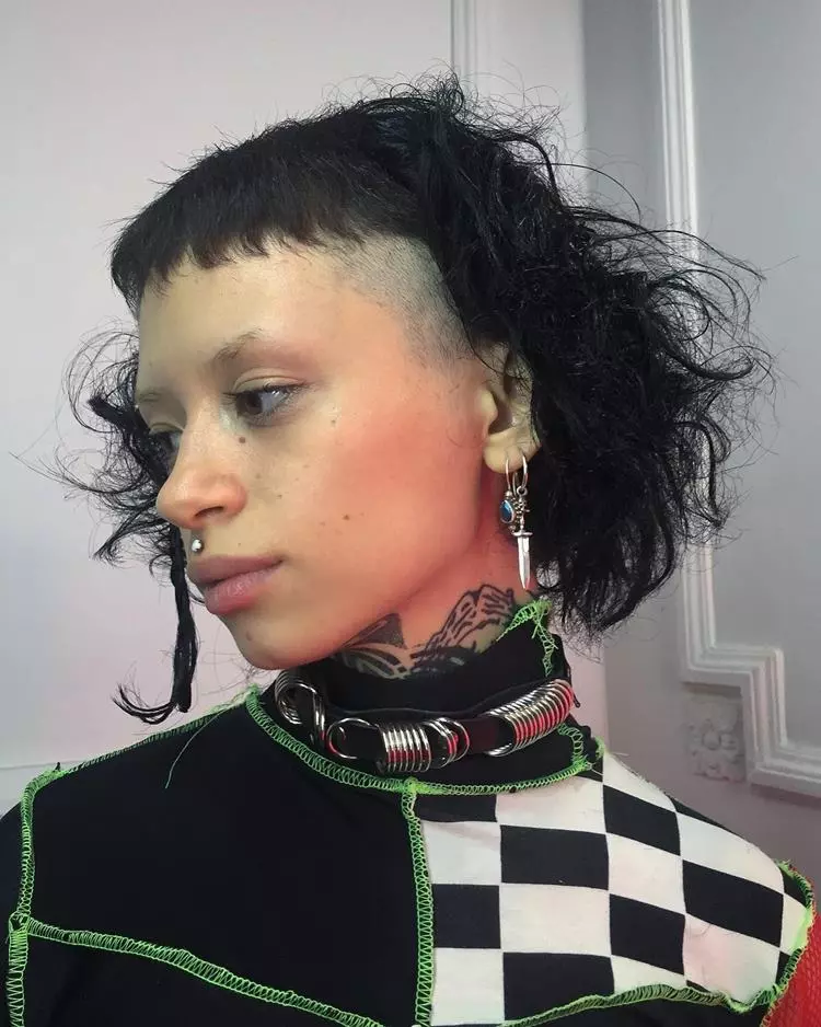 Menina com corte de cabelo curto com microfranja, usando piercings e joias prateadas de espada. Sua roupa é preta feita de retalhos