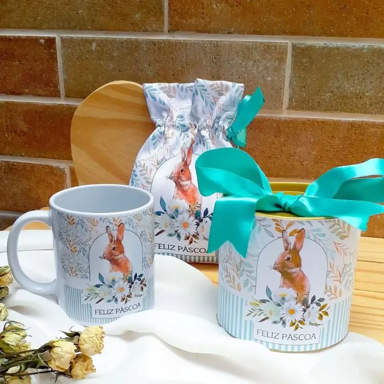 Conjunto decorativo de Páscoa com estampa de coelho: caneca, caixa com laço azul e sacolinha temática adornada com flores. Fundo com tijolos e mesa clara. Tons suaves e detalhes florais evocam delicadeza e celebração da data especial.