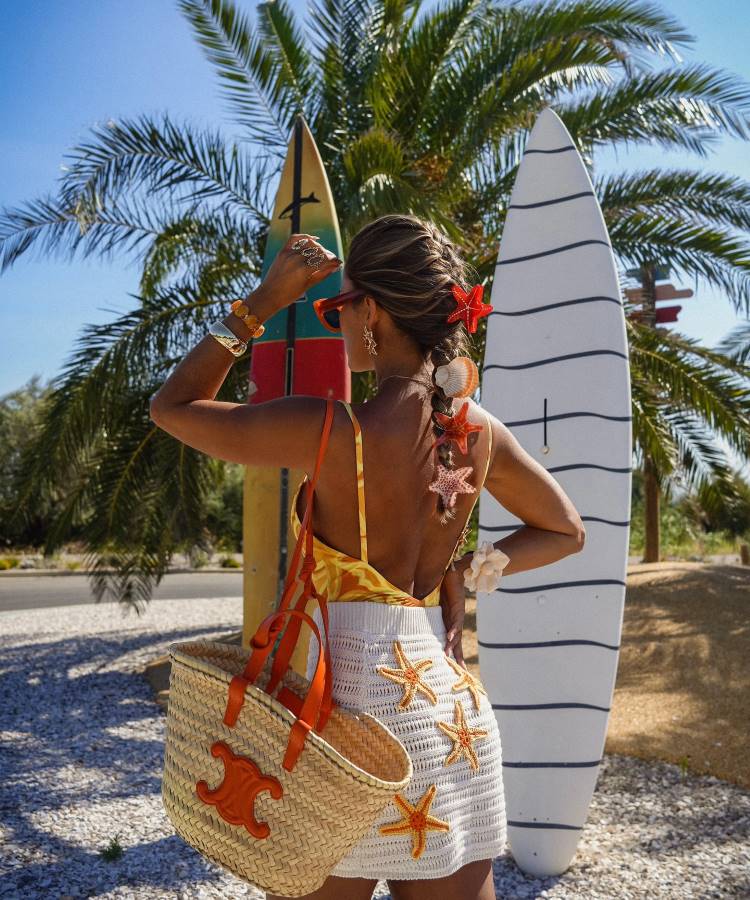 Uma mulher de costas, com tranças decoradas com estrelas-do-mar, posa ao lado de duas pranchas de surfe. Ela usa um maiô amarelo e um shorts de crochê branco com estrelas-do-mar e carrega uma bolsa de palha grande.
