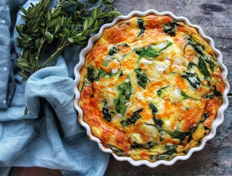 Prato com quiche de queijo e espinafre, receita de brunch de Dia das Mães, sobreposto em superfície de madeira ao lado de tecido e ramo de folhas verdes.