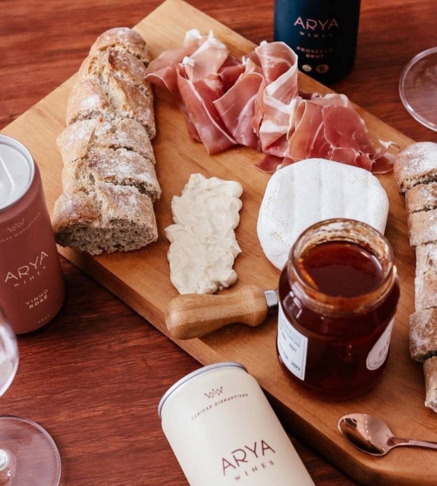 tábua com pães, queijos, presunto cru e geleia ao lato de latas de vinho