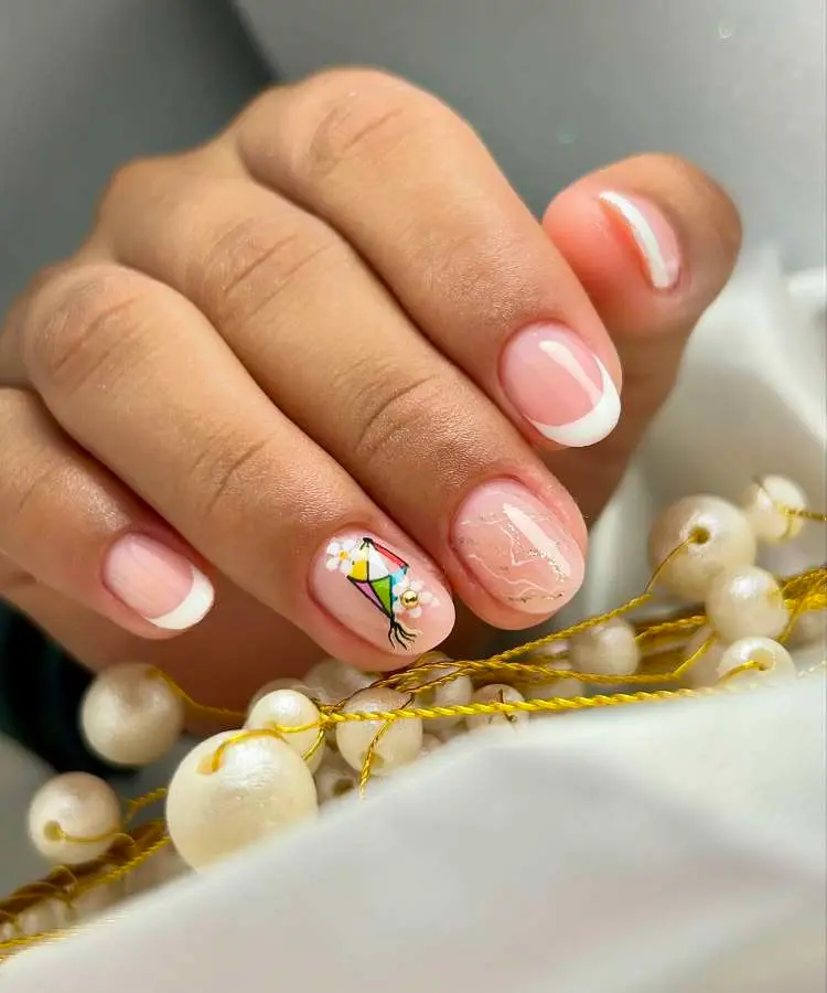 manicure com unhas curtas e arredondadas. Duas unhas possuem francesinha clássica branca. Uma unha se destaca com um delicado desenho de pipa e flores, enquanto outra tem um brilho sutil e linhas douradas finas, lembrando um bracelete.