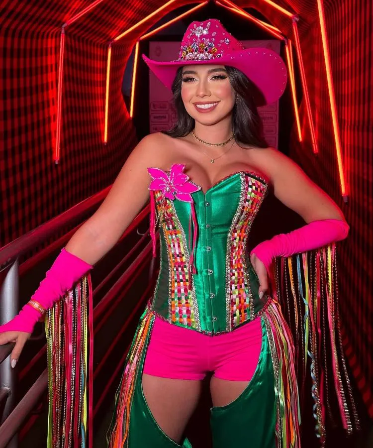 Mulher em chapéu rosa brilhante, cabelo ondulado, maquiagem marcante. Veste corset verde com detalhes coloridos e shorts rosa. Mangas com franjas multicoloridas. Fundo iluminado em vermelho com padrão xadrez e luzes de neon. Atmosfera vibrante e festiva.
