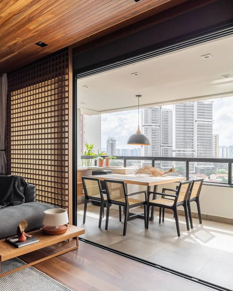 Varanda do apartamento decorado de 187 m². Mesa de 4 cadeiras, com luminária preta e bronze. De decoração no centro, um tronco enraizado seco.