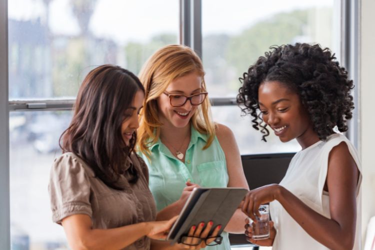 Três mulheres sorridentes, de diversas etnias, colaboram em um tablet em um escritório iluminado. A imagem transmite trabalho em equipe e inovação, com uma mulher de cabelo escuro à esquerda, uma ruiva de óculos no centro e uma mulher negra com cabelo encaracolado à direita, todas engajadas na tecnologia.