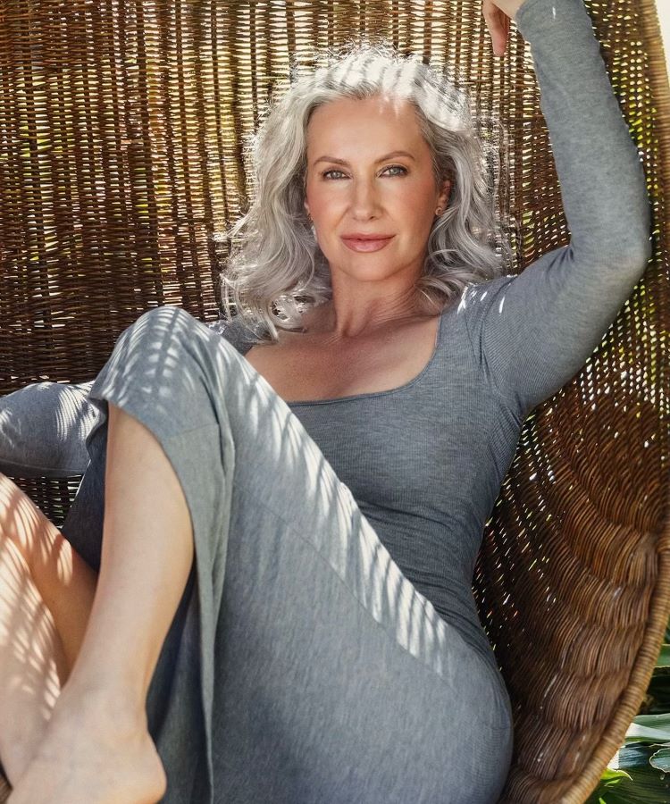 Mulher com cabelos grisalhos volumosos sentada em uma cadeira de vime ao ar livre. Ela usa um vestido cinza ajustado, com luzes e sombras projetadas pelas folhas ao redor, criando um efeito natural e relaxante.