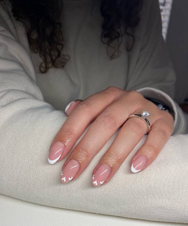 Mão feminina com unhas ovais decoradas com esmalte nude e design francês branco. Detalhes delicados de pequenos corações brancos adicionam charme. Um anel prateado com pedra brilhante enfeita o dedo, enquanto o fundo neutro destaca o estilo romântico.