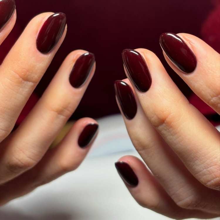 Mãos femininas com unhas no formato amendoado, pintadas na cor vermelho cereja intenso, destacando brilho e sofisticação. O fundo desfocado em tons de vermelho complementa a elegância do esmalte, inspirado nas tendências vibrantes de 2025.