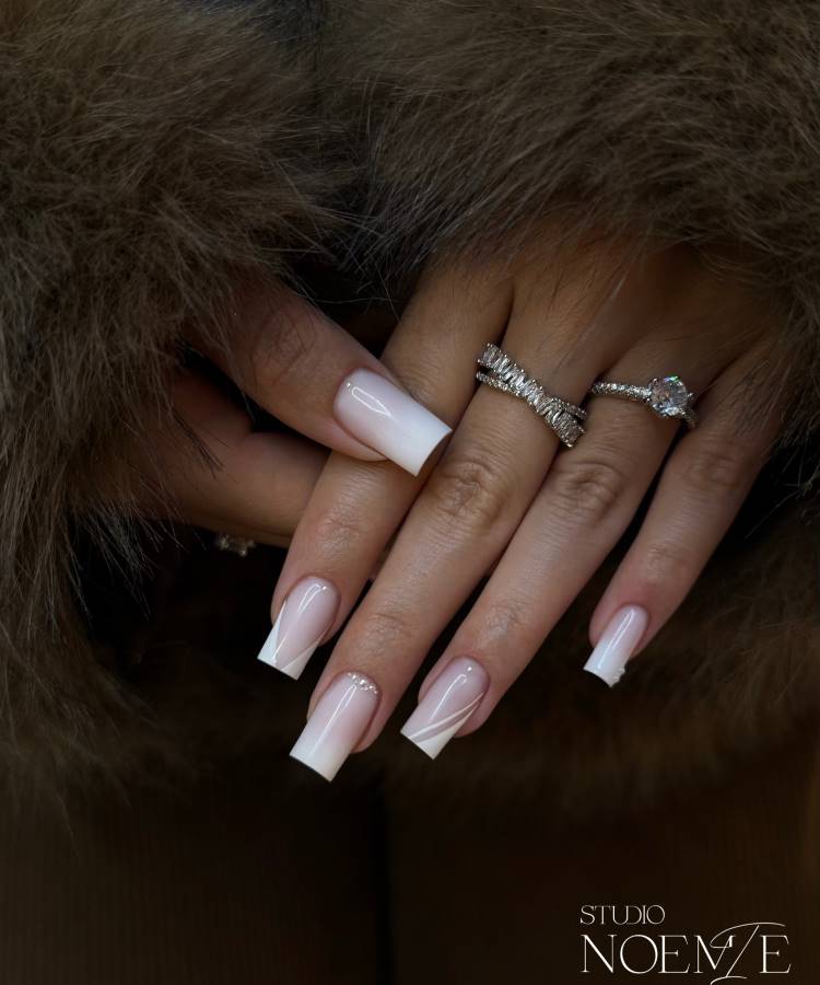 Mãos femininas com unhas em formato quadrado e design francês moderno, combinando nude e branco com um toque de arte geométrica. Anéis prateados detalhados e um fundo com textura de pelagem criam um visual sofisticado e luxuoso.





