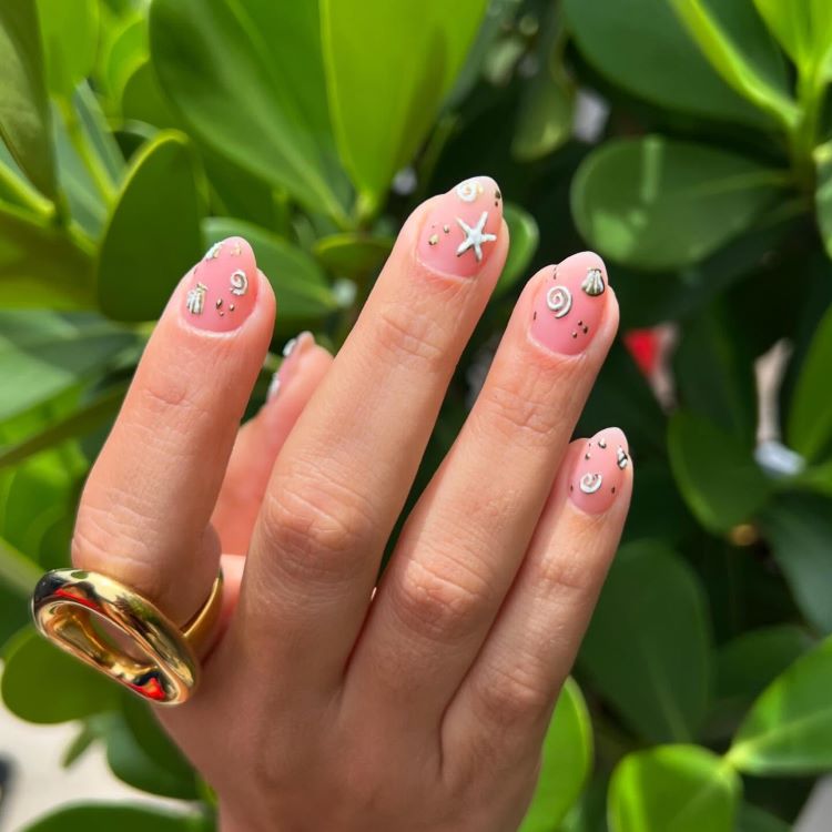 Mão destacando estilos de unhas curtas, decoradas com esmalte rosa claro e elementos marítimos como estrela-do-mar, conchas e detalhes prateados brilhantes. Dedos adornados com anel dourado grosso. Fundo com folhas verdes vibrantes, criando um contraste tropical e delicado.