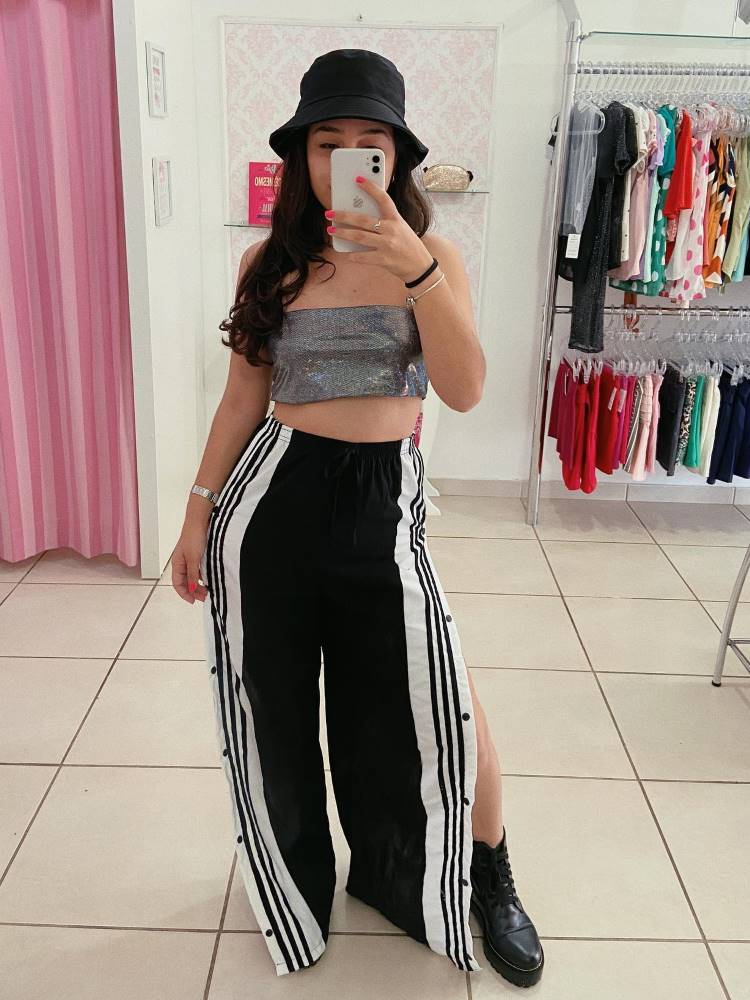 Mulher de pele clara e cabelo preto usando top faixa holográfico, calça preta e branca aberta nas laterias e bucket