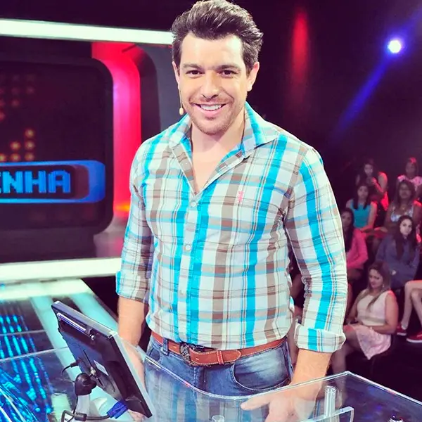 Cézar usando camisa xadrez marrom e azul e calça jeans no palco do Mega Senha