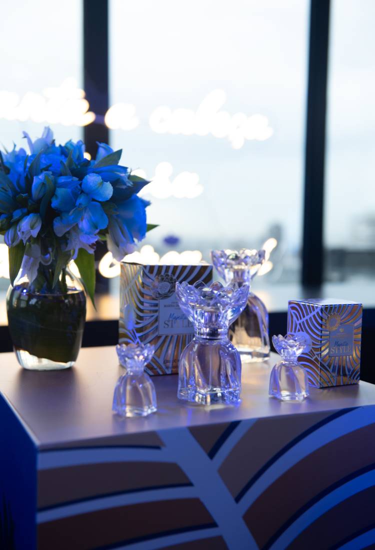 Frascos de perfume com design decorativo sobre mesa com padrão azul e dourado. Embalagens correspondentes ao fundo. Vaso de flores azuis ao lado. A luz ambiente cria uma atmosfera elegante, destacando os produtos e a decoração sofisticada da mesa.