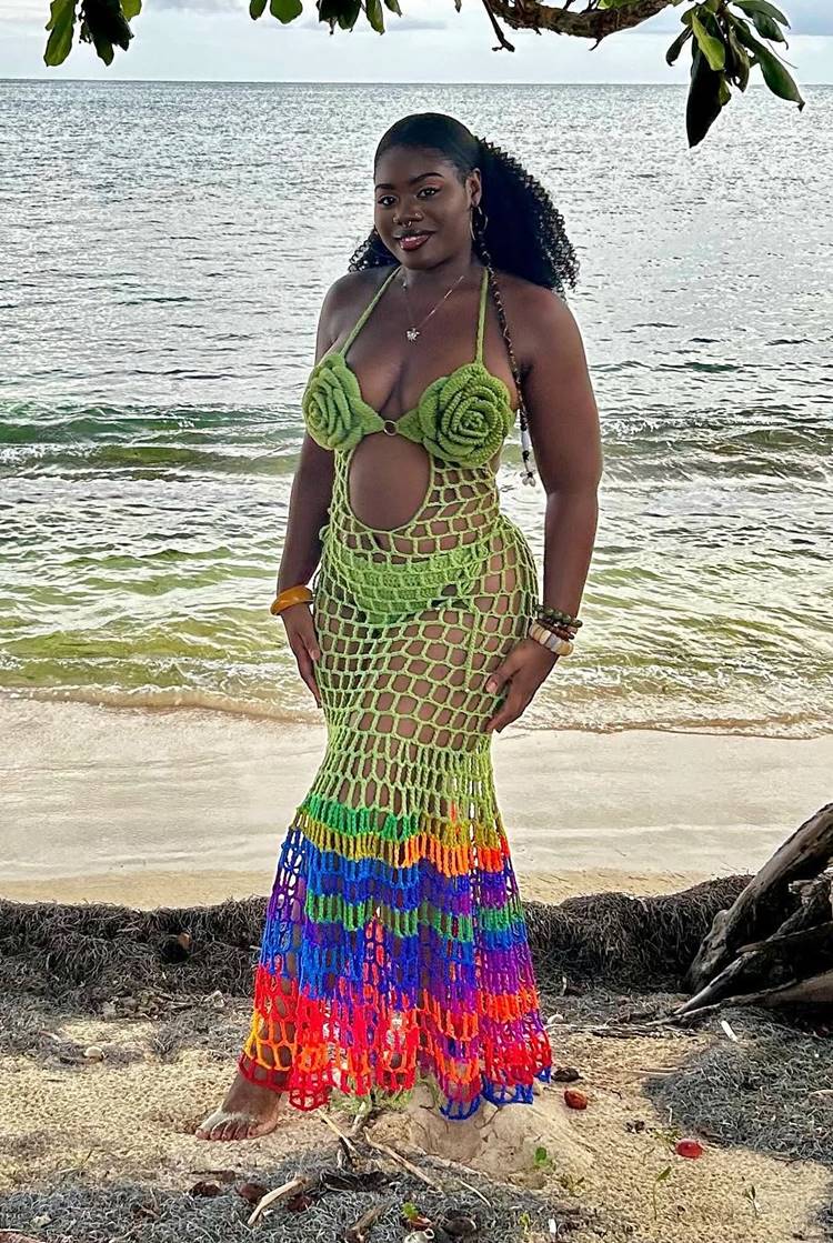 Uma mulher negra sorri na praia, vestindo um vestido de crochê verde e colorido. O top tem flores de crochê e a saia longa apresenta um padrão de rede vazada e listras nas cores do arco-íris na barra.