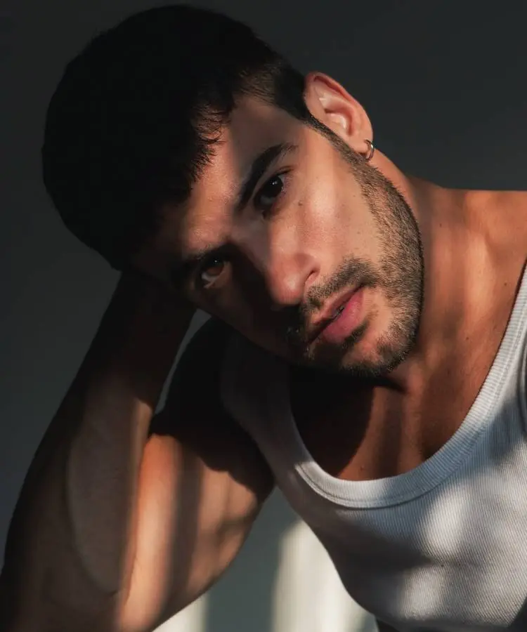 Homem com barba e cabelos escuros, vestindo uma regata branca. Ele olha para a câmera com uma expressão introspectiva. A luz do sol da tarde projeta sombras em listras em seu braço e ombro.