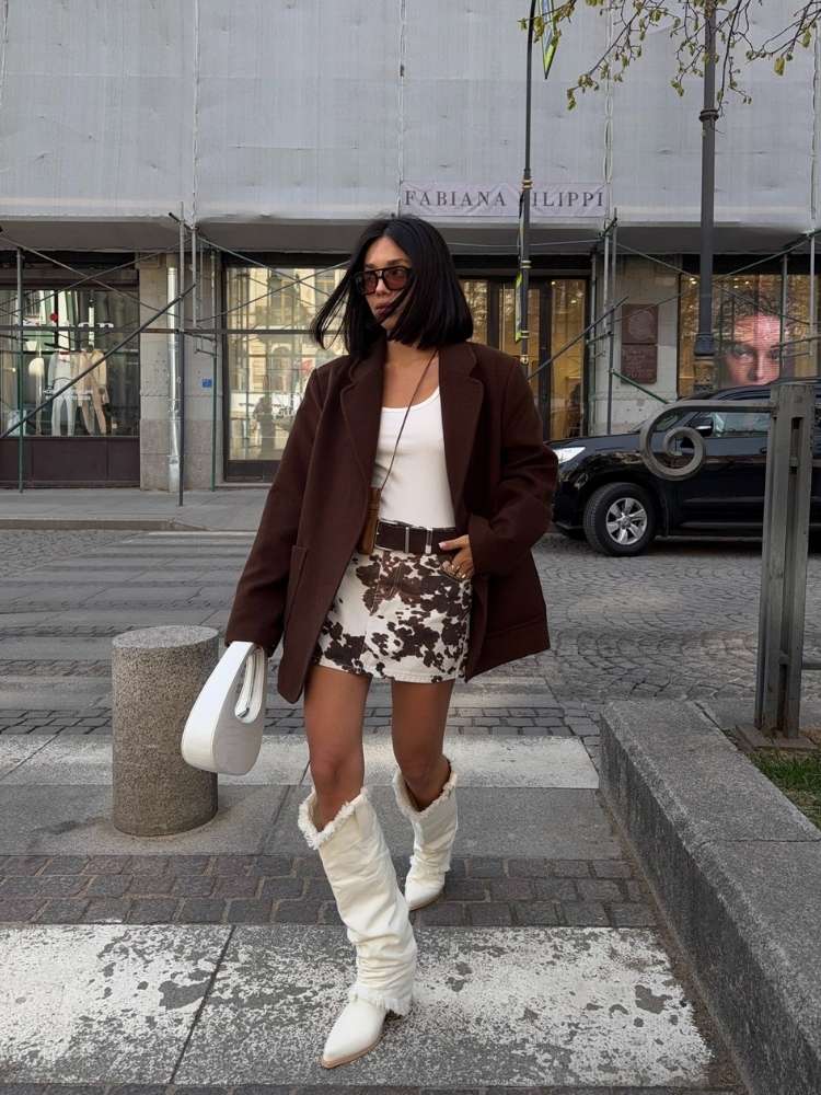 Mulher de pele clara de regata branca, blazer marrom, saia cow print e bota western branca 
